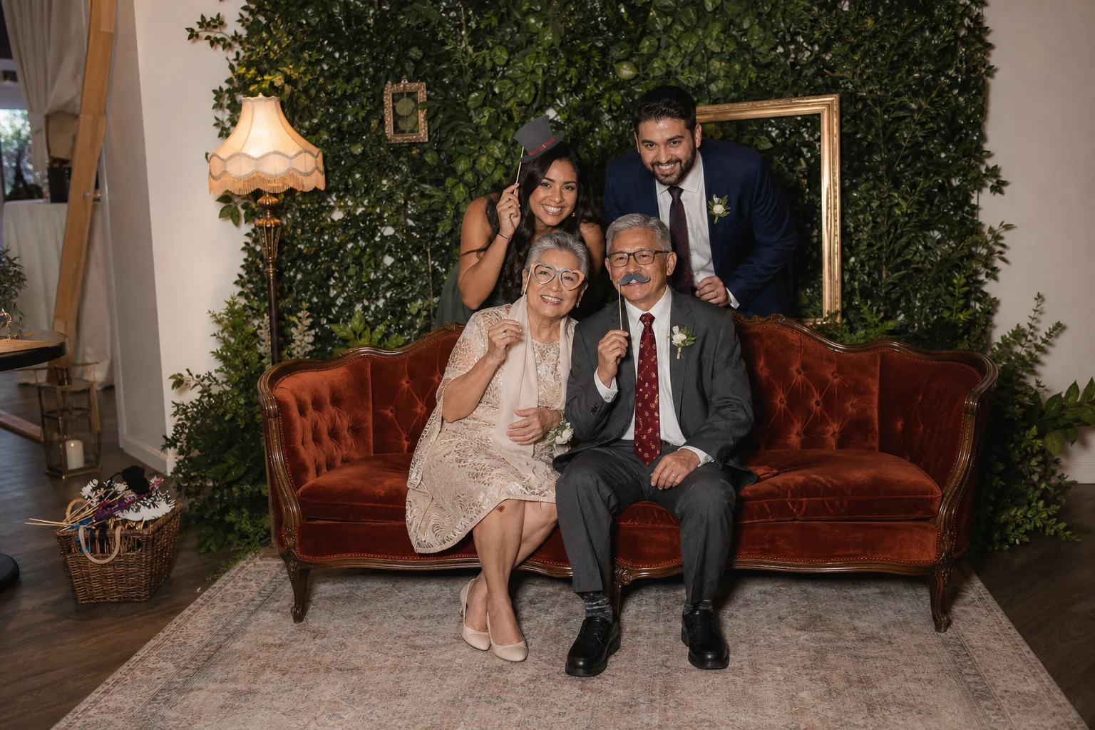 Wedding guests posing together on a rust velvet sofa in front of a greenery photo booth backdrop